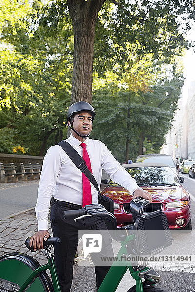 Thoughtful businessman standing by bicycle against tree