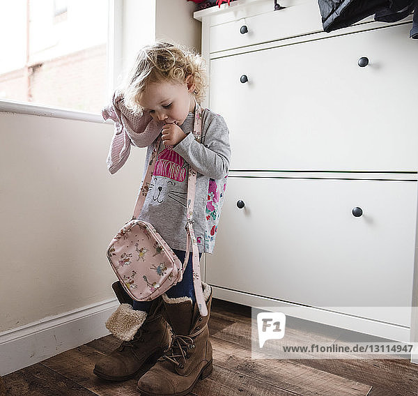 Girl in big shoes licking thumb while standing by window at home