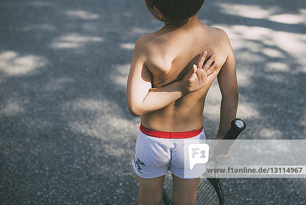 Rückansicht eines Jungen ohne Hemd  der auf der Straße einen Tennisschläger hält