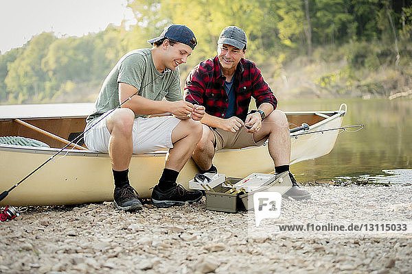 Smiling friends talking while adjusting fishing tackles on boat at lakeshore