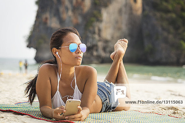 Frau hört Musik über Smartphone  während sie am Strand auf einer Matte liegt