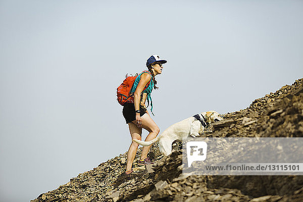 Side view of woman carrying backpack with dog climbing mountain against clear sky
