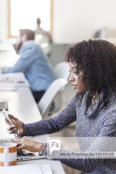 Geschäftsfrau benutzt Telefon und Laptop  während sie im Kreativbüro am Schreibtisch sitzt
