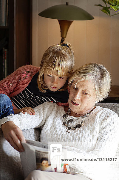 Grandmother showing magazine to granddaughter while sitting on sofa at home