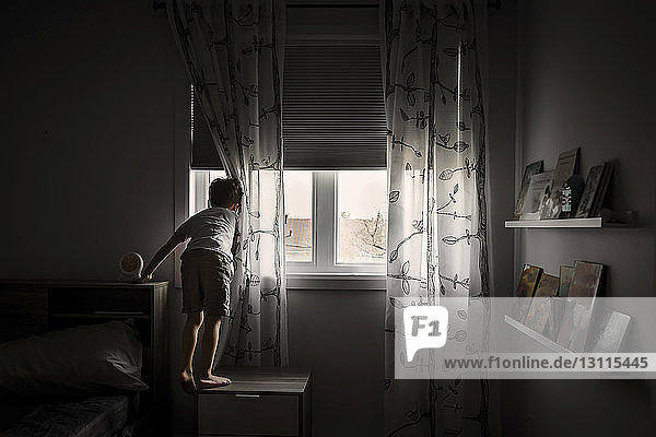 Rückansicht eines Jungen  der durch ein Fenster schaut  während er zu Hause auf einem Schrank steht