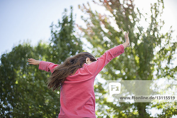 Niedrigwinkelansicht eines verspielten Mädchens mit ausgestreckten Armen an Bäumen im Garten