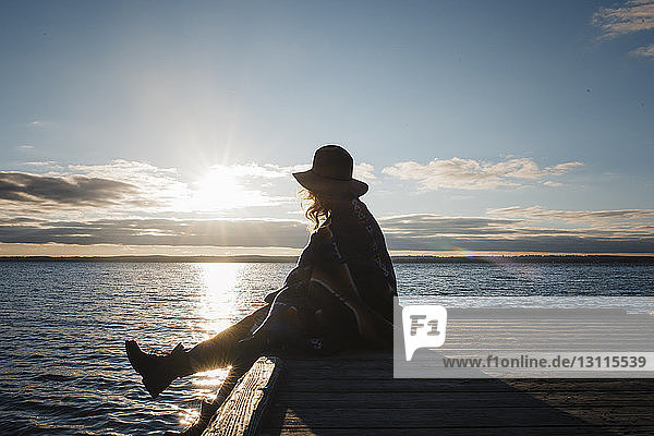 Side view of woman wrapped in blanket sitting on pier by Lake Simcoe during sunset