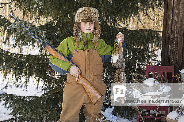 Portrait of smiling boy holding hare and rifle while standing in backyard