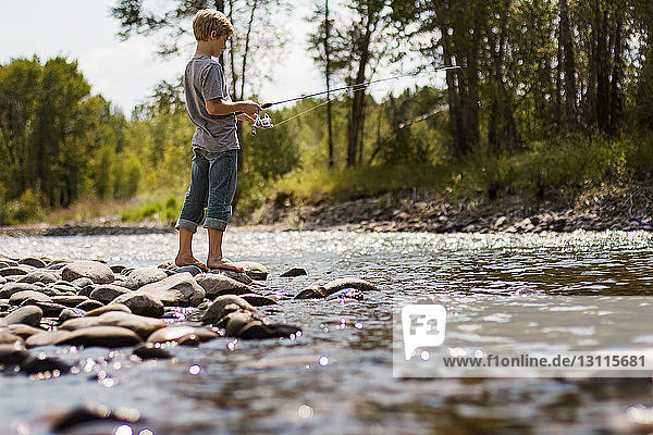 Seitenansicht eines Jungen  der auf Felsen stehend im Fluss im Wald fischt