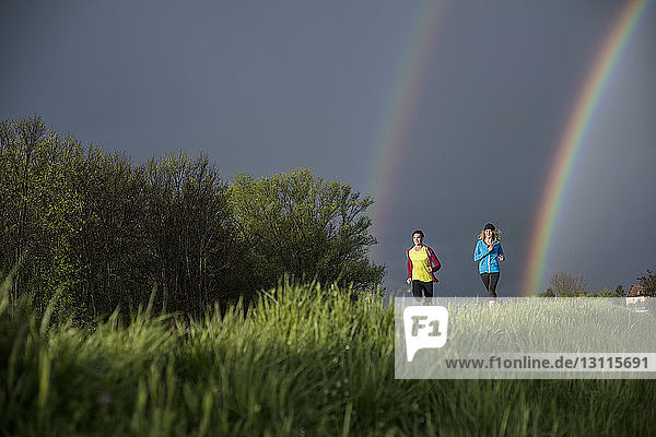 Freunde laufen auf Grasfeld gegen den Himmel mit doppeltem Regenbogen