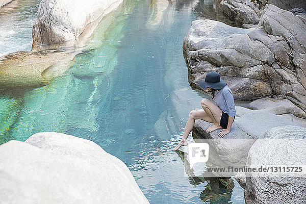Seitenansicht einer Wanderin  die auf einem Felsen sitzend ihren Fuß in den Fluss taucht