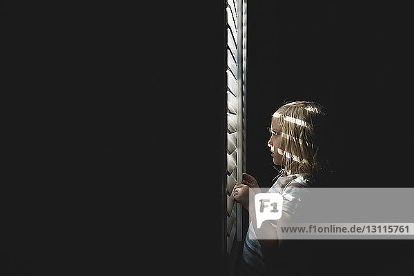 Side view of girl looking through blinds on sunny day