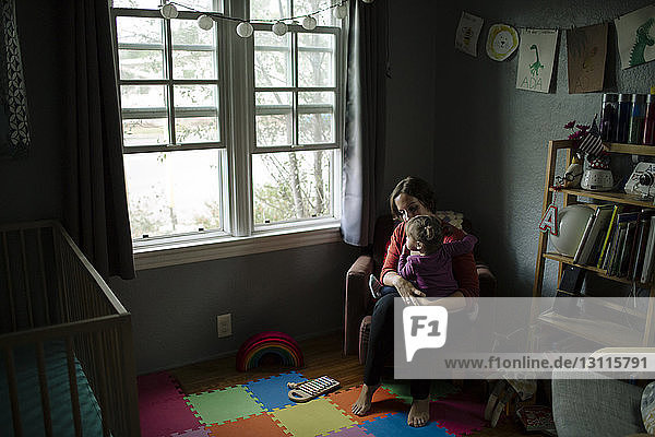 Hochwinkelaufnahme einer Tochter  die durch ein Fenster schaut  während sie zu Hause auf dem Schoß der Mutter sitzt