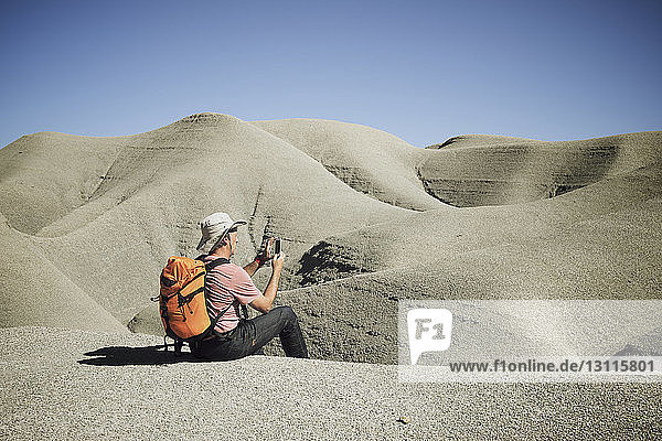 Rucksacktourist fotografiert per Handy  während er im Sand der Wüste vor klarem Himmel sitzt