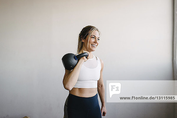 Smiling female athlete carrying kettlebell while standing by wall in gym