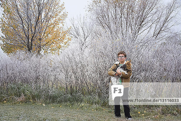 Porträt eines Jungen  der einen Hund trägt  während er im Winter auf einem Grasfeld im Park steht