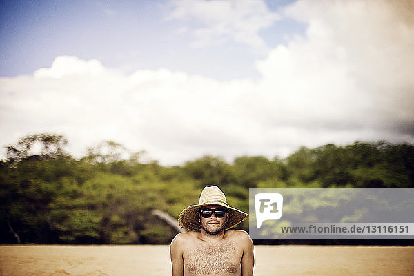 Porträt eines Mannes mit Strohhut  der sich am Strand vor bewölktem Himmel entspannt