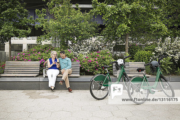 Älteres Paar  das ein Smartphone benutzt und auf einer Bank am Fußweg im Park sitzt