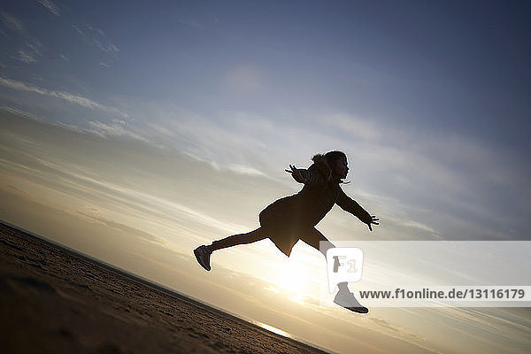 Glückliches Mädchen springt bei Sonnenuntergang am Strand gegen den Himmel