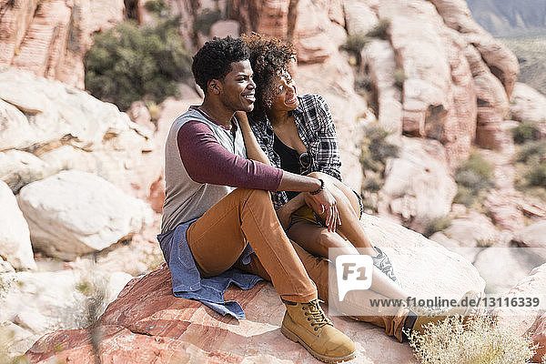 Couple looking away while sitting on rock formation during sunny day
