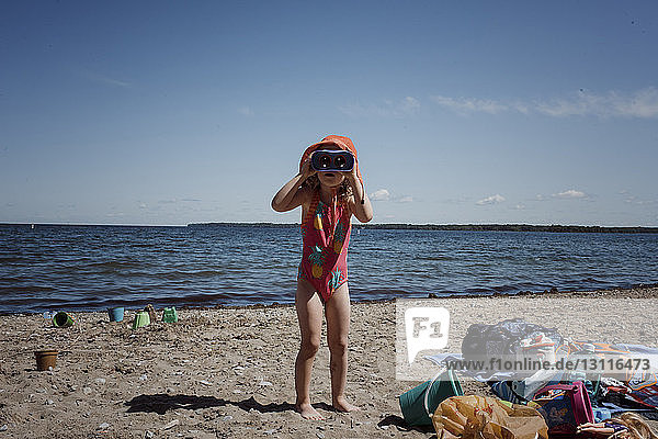 Mädchen in Badebekleidung schaut durch ein Fernglas  während sie am Strand vor blauem Himmel am sonnigen Tag steht