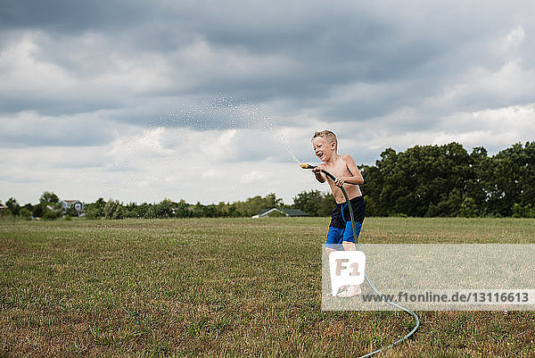 Full length of shirtless playful boy playing with garden hose on grassy field against cloudy sky at backyard