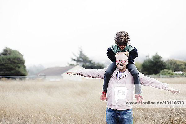 Playful father with arms outstretched carrying son on shoulders at field against clear sky