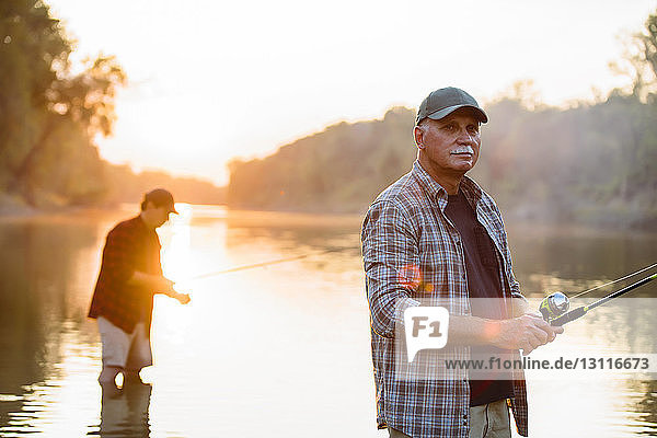Senior man looking away while friend fishing in background during sunset