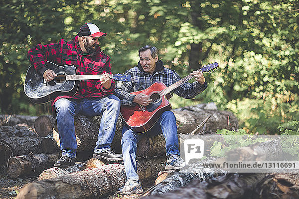 Friends playing guitars while sitting on logs in forest