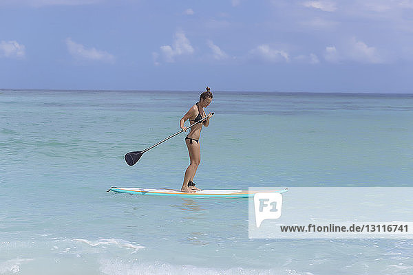 Frau im Bikini paddelt bei Sonnenschein auf dem Meer gegen den Himmel