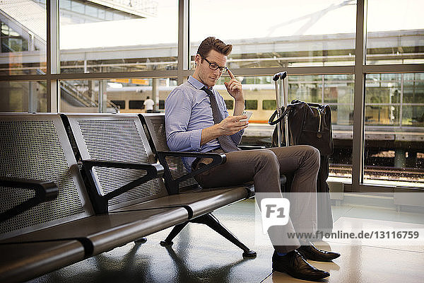 Businessman using smart phone while sitting on seat in waiting room at railroad station