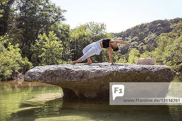 Frau praktiziert Yoga auf dem Gipfel eines Felsens inmitten eines Sees im Wald