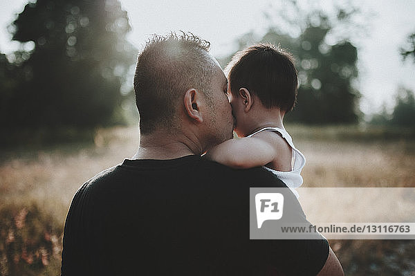 Rückansicht eines Vaters  der seinen Sohn küsst  während er auf einem Grasfeld steht