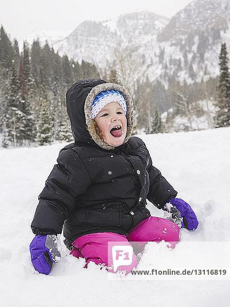 Happy baby girl kneeling on snow covered field against mountain