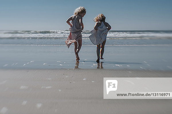 Rückansicht von Schwestern  die beim Strandspaziergang bei klarem Himmel ein Kleid in der Hand halten