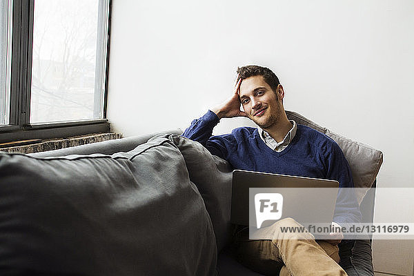 Lächelnder Mann benutzt Laptop  während er zu Hause auf dem Sofa sitzt