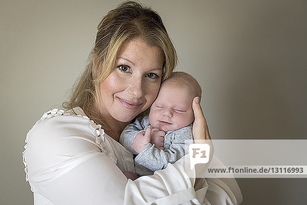 Portrait of confident mother carrying cute sleeping son against wall at home