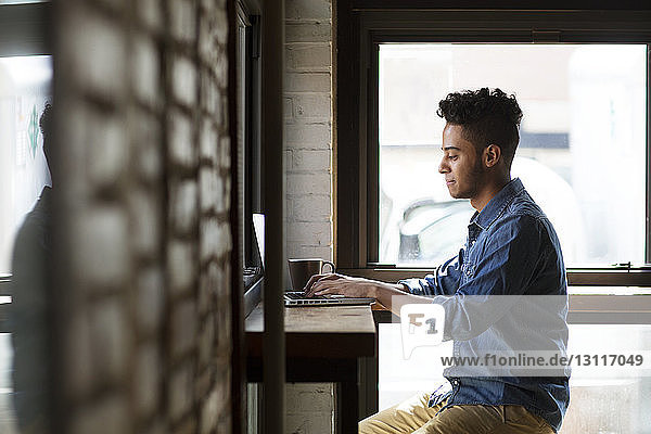 Seitenansicht eines Mannes  der einen Laptop benutzt  während er an den Fenstern eines Cafés sitzt