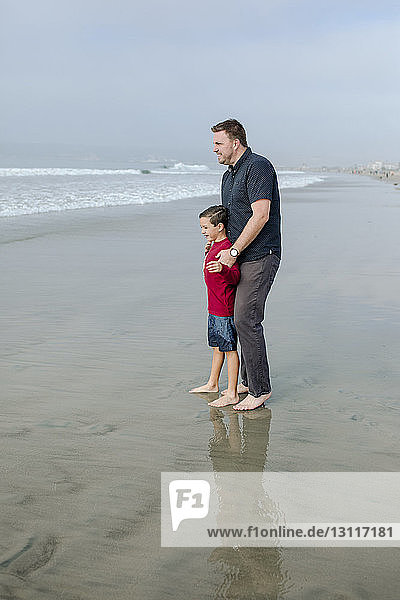Full length of father and son looking at view while standing on shore at beach