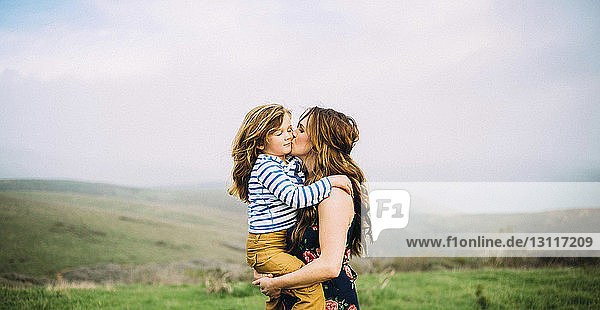 Loving woman kissing daughter while standing on field against sky