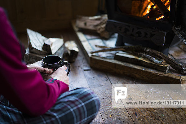 Midsection of man having drink while sitting by wood burning stove in cottage