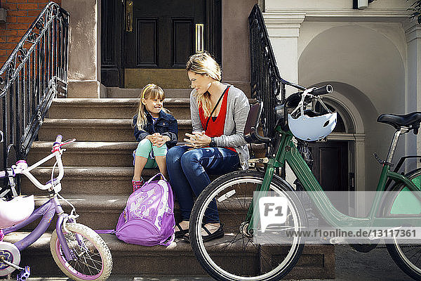 Mother and daughter talking while sitting on steps with bicycles parked in foreground
