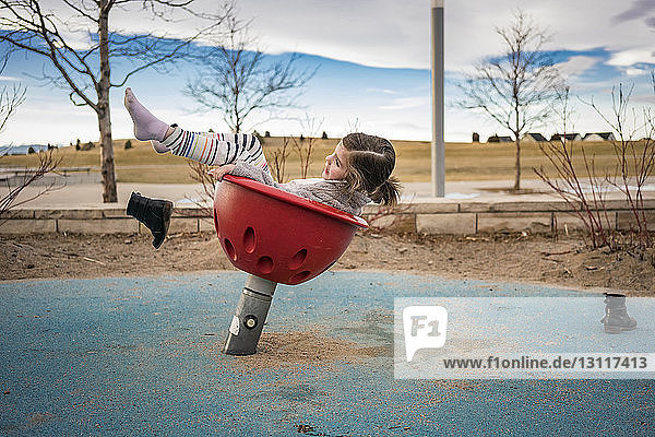 Seitenansicht eines verspielten Mädchens  das Schuhe wirft  während es auf einem Spielgerät im Freien gegen den bewölkten Himmel im Park spielt