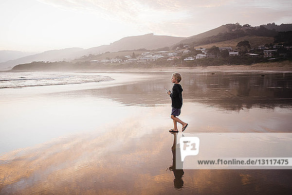 Side view of boy looking away while walking at beach against sky during sunset
