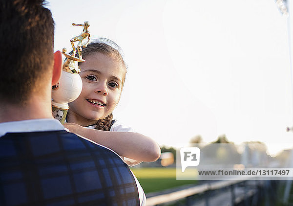 Rear view of father carrying daughter holding trophy at park against clear sky during sunset