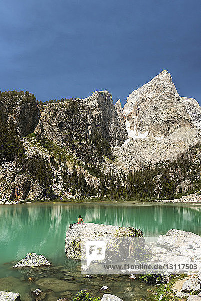 Distanzansicht eines Teenagers  der auf einem Felsen am See vor Bergen sitzt