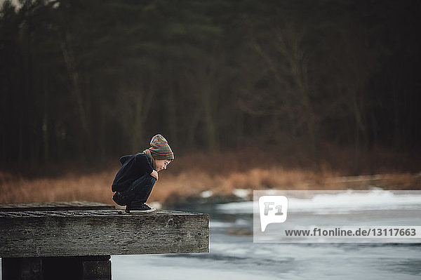 Seitenansicht eines neugierigen Jungen  der auf den Fluss schaut  während er im Park auf dem Holz kauert