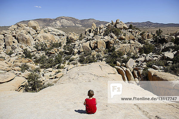 Rear view of boy looking at rock formations while sitting at Joshua Tree National Park against sky