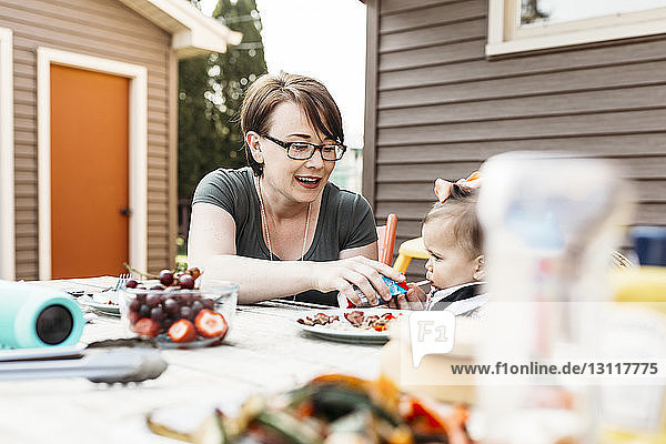 Mother feeding daughter while sitting at table in backyard