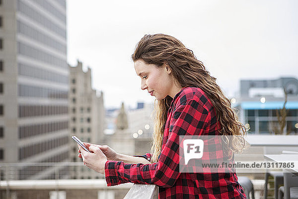Seitenansicht einer Frau  die ein Smartphone benutzt  während sie sich am Geländer eines Terrassencafés anlehnt